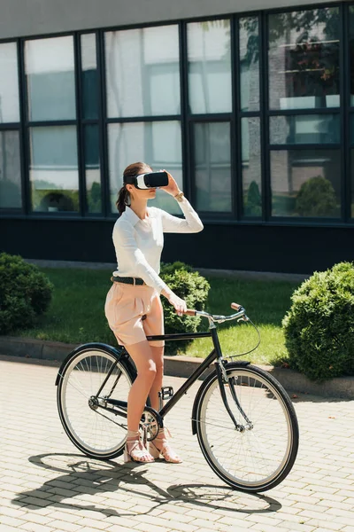 Businesswoman touching virtual reality headset and sitting on bicycle near building — Stock Photo