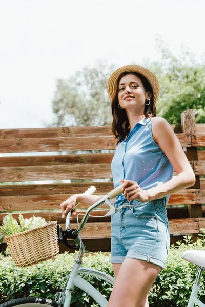 Cheerful girl in straw hat standing with bicycle outside — Stock Photo