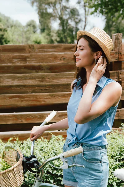 Cheerful girl in straw hat touching hair and standing with bicycle near fence — Stock Photo