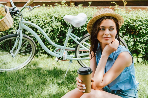 Дівчина в солом'яному капелюсі тримає паперову чашку і сидить біля велосипеда — стокове фото