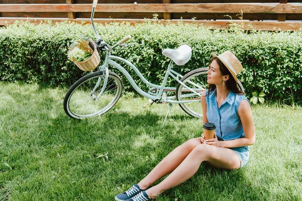 Femme en chapeau de paille tenant tasse en papier et assis sur l'herbe près du vélo — Photo de stock