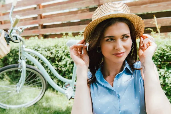Привлекательная женщина трогает соломенную шляпу рядом с велосипедом и забором — стоковое фото
