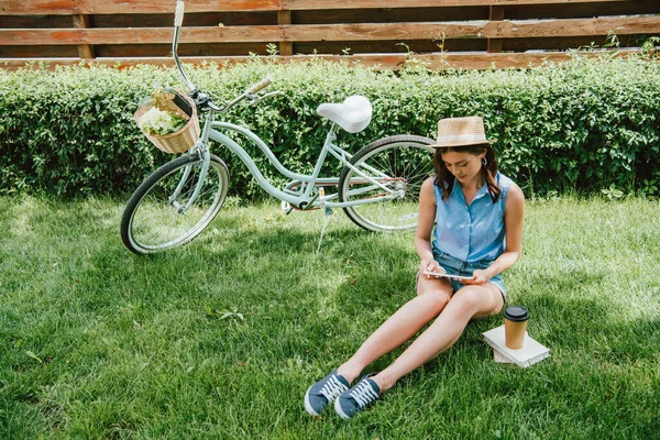Chica en sombrero de paja usando tableta digital y sentado en la hierba cerca de la bicicleta, taza de papel y libros - foto de stock