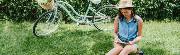 Concept panoramique de fille en chapeau de paille en utilisant une tablette numérique tout en étant assis sur l'herbe près de vélo — Photo de stock