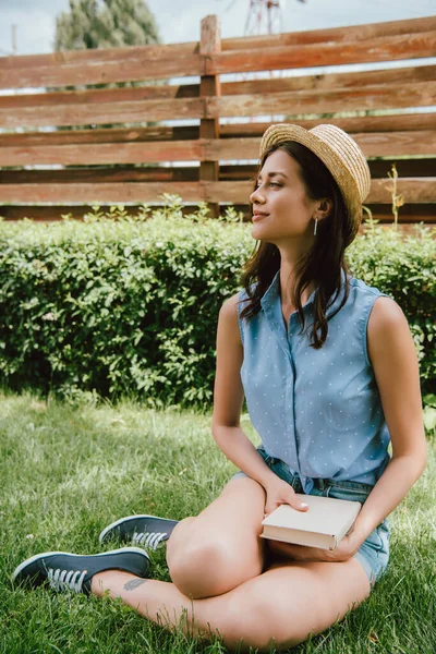 Happy girl in straw hat holding book and sitting on grass — Stock Photo