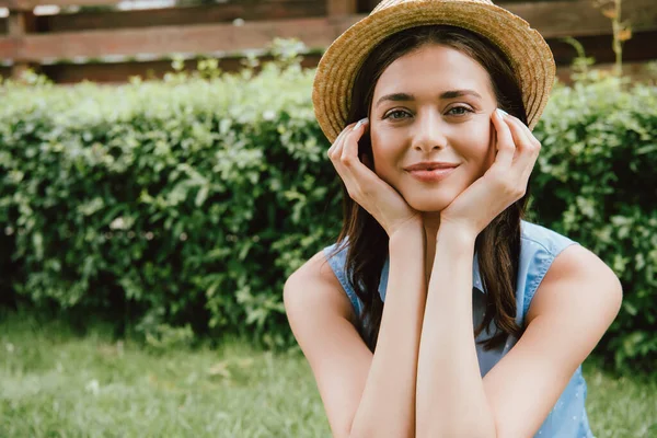 Cheerful young woman in straw hat touching face and looking at camera — Stock Photo
