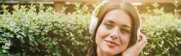 Panoramic shot of cheerful young woman listening music in wireless headphones near bush — Stock Photo