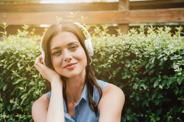 Mujer feliz escuchando música en auriculares inalámbricos cerca de arbusto - foto de stock