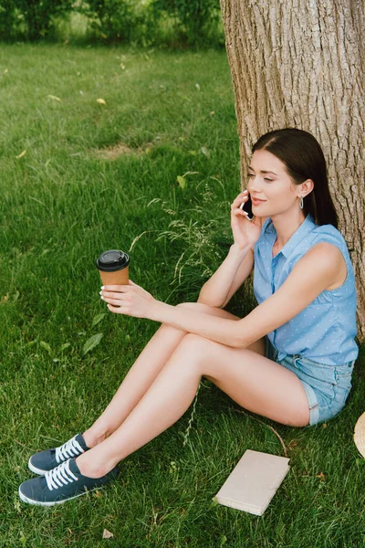 Hermosa mujer hablando en el teléfono inteligente y sosteniendo la taza de papel mientras está sentado en la hierba - foto de stock