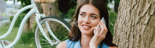 Foto panorámica de la mujer feliz hablando en el teléfono inteligente cerca de la bicicleta - foto de stock