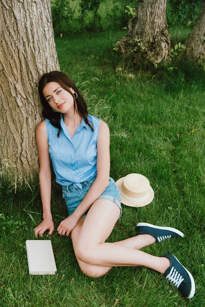 Donna felice seduta sull'erba vicino tronchi d'albero, libro e cappello di paglia — Foto stock