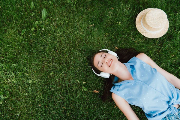 Draufsicht einer fröhlichen Frau, die im Gras liegt und neben Strohhut Musik hört — Stockfoto