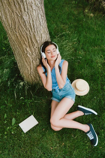 Hochwinkel-Ansicht der schönen Frau mit geschlossenen Augen Musik hören, während sie auf Gras in der Nähe von Buch und Strohhut sitzt — Stockfoto