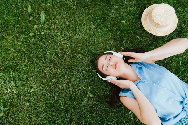 Vista superior de la mujer bonita con los ojos cerrados acostado en la hierba y escuchar música cerca de sombrero de paja - foto de stock