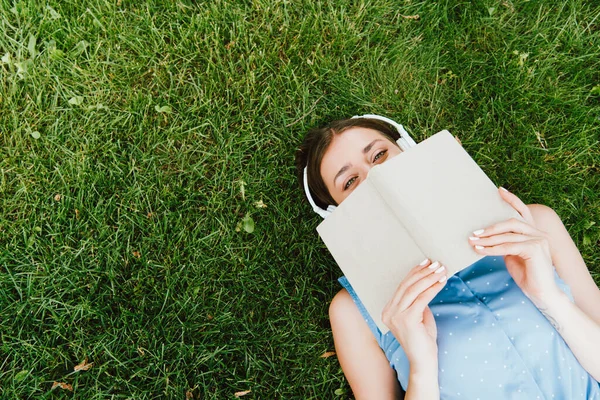 Draufsicht einer Frau mit drahtlosen Kopfhörern, die im Gras liegt und Buch in der Hand hält — Stockfoto