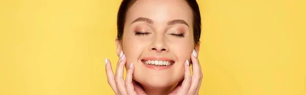 Feliz hermosa mujer con los ojos cerrados tocando la cara aislada en amarillo, plano panorámico - foto de stock