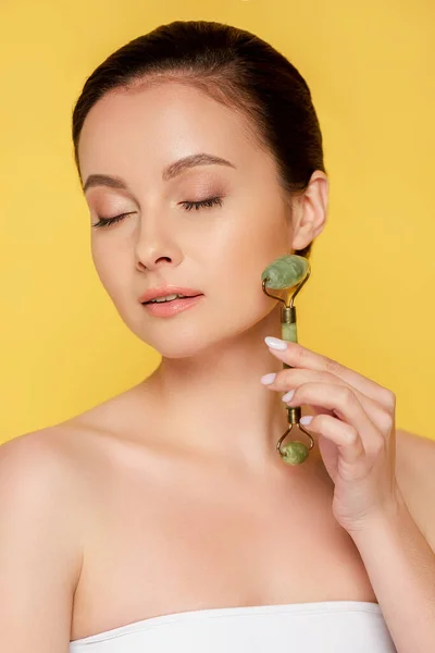 Beautiful woman with closed eyes using jade roller on face isolated on yellow — Stock Photo