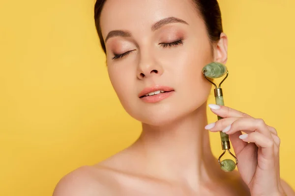 Beautiful naked woman with closed eyes using jade roller on face isolated on yellow — Stock Photo