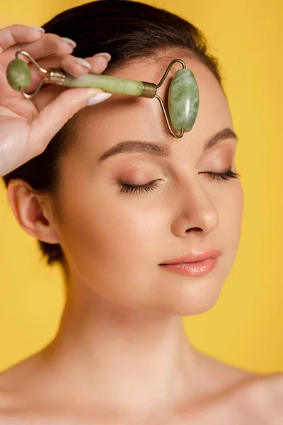 Belle femme nue avec les yeux fermés à l'aide de rouleau de jade sur le visage isolé sur jaune — Photo de stock