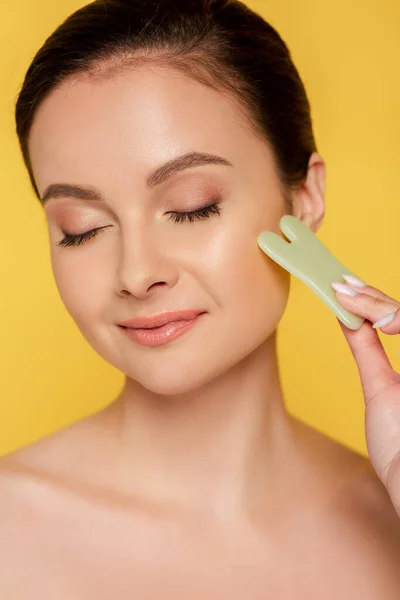 Retrato de hermosa mujer desnuda con los ojos cerrados facial jade gua sha aislado en amarillo - foto de stock