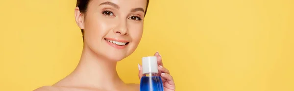 Sonriente hermosa mujer desnuda sosteniendo agua micelar en botella aislada en amarillo, tiro panorámico - foto de stock