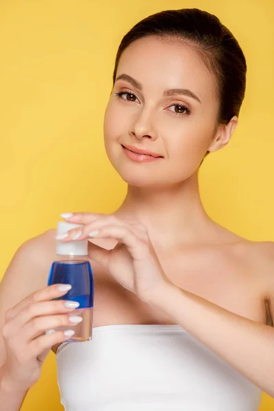 Smiling beautiful woman holding micellar water in bottle isolated on yellow — Stock Photo