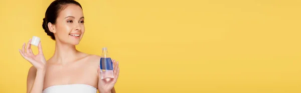 Sonriente hermosa mujer sosteniendo micelar agua en botella aislado en amarillo, tiro panorámico - foto de stock