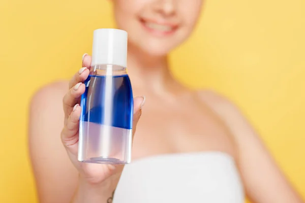 Enfoque selectivo de sonriente hermosa mujer sosteniendo agua micelar en botella aislada en amarillo - foto de stock