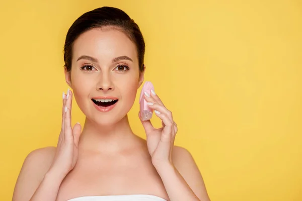 Shocked beautiful woman holding facial cleansing brush isolated on yellow — Stock Photo