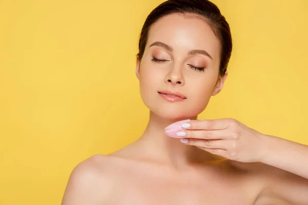 Beautiful naked woman using facial cleansing brush with closed eyes isolated on yellow — Stock Photo