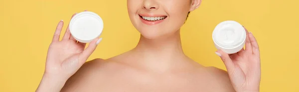 Vista cortada de sorrir bela mulher nua segurando recipiente com creme cosmético isolado em amarelo, tiro panorâmico — Fotografia de Stock