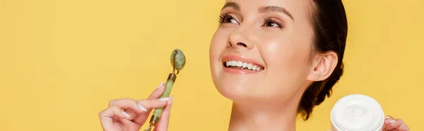 Happy beautiful woman holding cosmetic cream and jade roller isolated on yellow, panoramic shot — Stock Photo