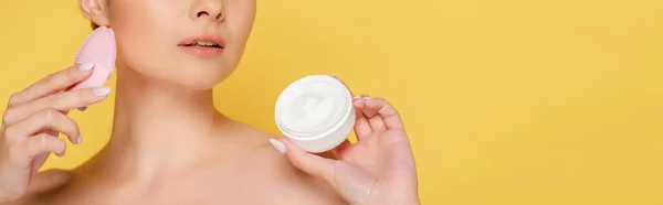 Cropped view of naked beautiful woman holding cosmetic cream and facial cleansing brush isolated on yellow, panoramic shot — Stock Photo