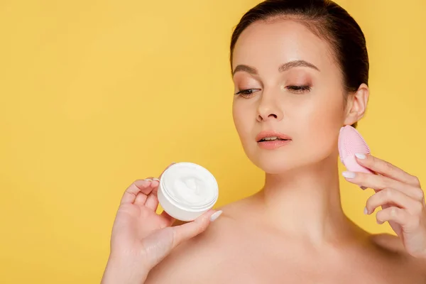 Hermosa mujer desnuda sosteniendo crema cosmética y cepillo de limpieza facial aislado en amarillo - foto de stock