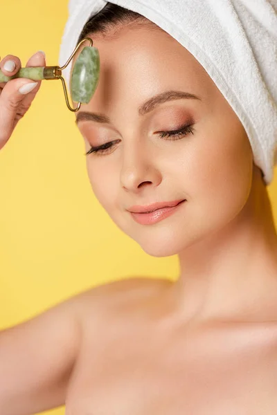 Bella donna nuda con asciugamano sulla testa utilizzando rullo di giada isolato su giallo — Foto stock