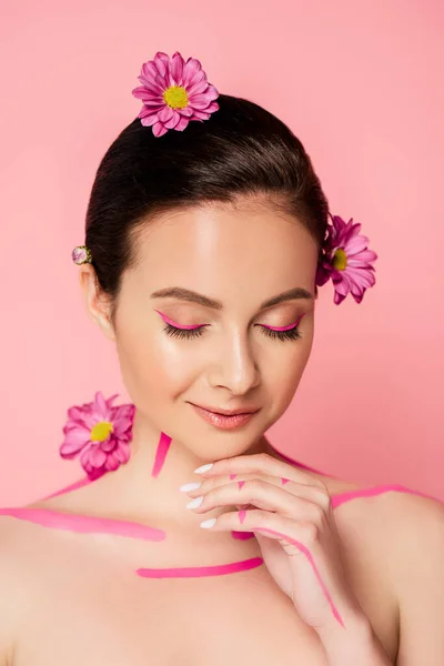 Hermosa mujer desnuda con los ojos cerrados, líneas de color rosa en el cuerpo y flores en el pelo aislado en rosa - foto de stock