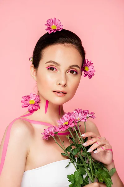 Hermosa mujer con líneas de color rosa en el cuerpo y flores en el pelo que sostiene ramo aislado en rosa - foto de stock