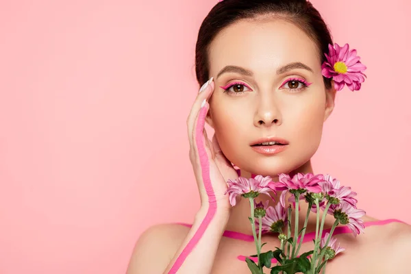 Naked beautiful woman with pink lines on body and flower in hair holding bouquet and touching face isolated on pink — Stock Photo