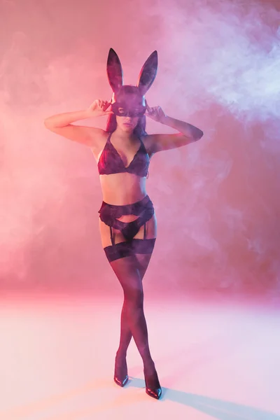 Sexy woman in underwear touching mask with bunny ears on pink with smoke — Stock Photo