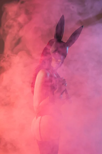 Mujer seductora en ropa interior sexy y máscara con orejas de conejo sosteniendo azotes látigo en rosa con humo - foto de stock
