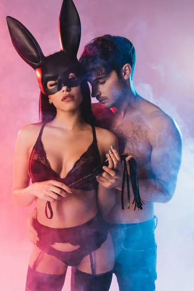 Мужчина без рубашки рядом с соблазнительной женщиной в маске с кроличьими ушами, держащими порку на розовом с дымом — стоковое фото