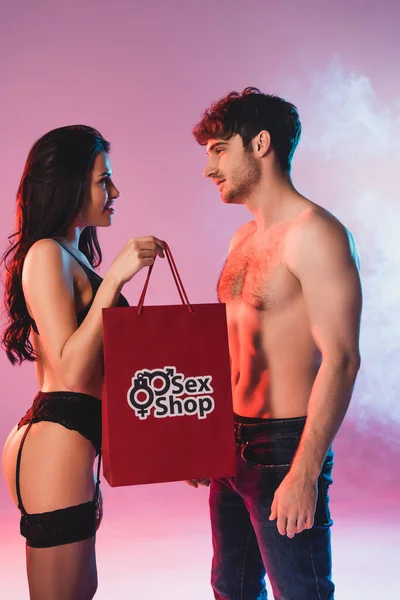 Vista lateral de la mujer sexy sosteniendo bolsa de compras con letras tienda de sexo y mirando al hombre sin camisa en rosa con humo - foto de stock