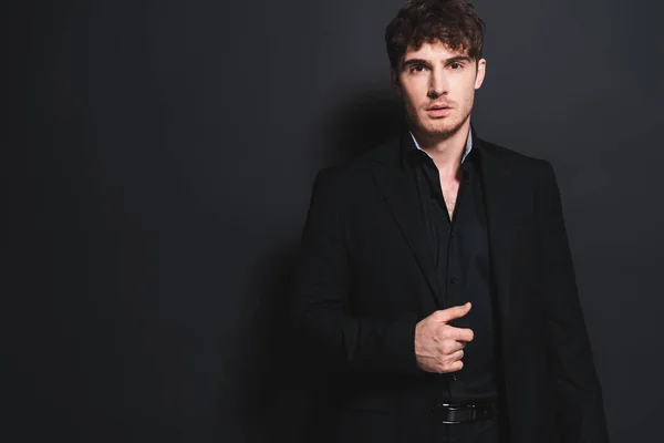 Hombre guapo en ropa formal tocando chaqueta y mirando a la cámara en negro - foto de stock