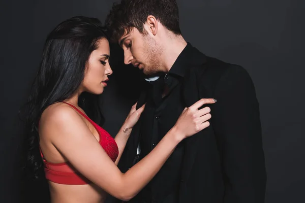 Vista laterale della donna sexy in reggiseno rosso e pizzo toccare camicia di bell'uomo su nero — Foto stock