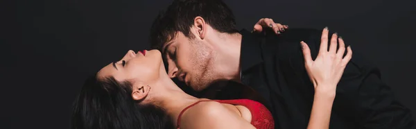 Panoramic crop of passionate man kissing neck of seductive woman in red bra isolated on black — Stock Photo