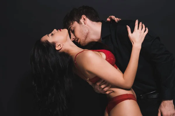 Appassionato uomo baciare collo di donna seducente in reggiseno rosso isolato su nero — Foto stock