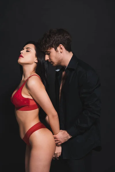 Homme passionné toucher les mains de la femme soumise en lingerie rouge isolé sur noir — Photo de stock