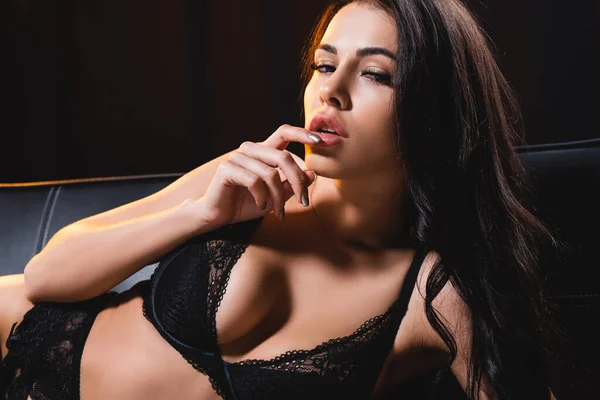 Mujer sexy en ropa interior de encaje acostado en sofá negro y tocando los labios - foto de stock