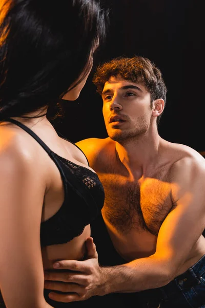 Uomo muscoloso toccare e guardare sexy donna in pizzo reggiseno isolato su nero — Foto stock