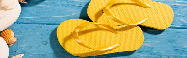 Draufsicht auf gelbe Flip Flops in der Nähe von Muscheln und Steinen auf blauem Holzgrund, Panoramaaufnahme — Stockfoto